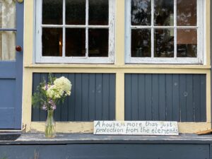 blue cottage with many windows and flowers in vase on ledge, sign with text "A house is more than just protection from the elements"